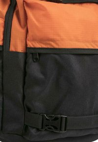 Svart ryggsekk med oransje toppseksjon, med teksturert stoff, glidelåslomme og plastspenne-lukking.