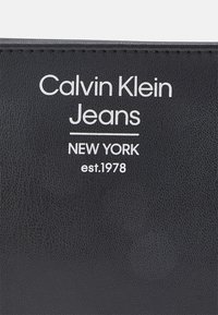 Černá kožená peněženka s vyraženým bílým textem: "Calvin Klein Jeans," "NEW YORK" a "est. 1978." Hladká textura s minimalistickým designem.