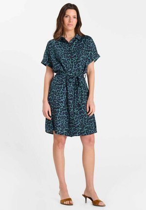 Femme portant une robe courte à manches courtes, longueur au genou, imprimé léopard bleu sarcelle et noir avec une ceinture à la taille, associée à des sandales à talons ouvertes marron.