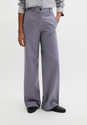 Personne portant un pantalon ample lavande avec poches latérales, associé à des mocassins noirs et une chemise à manches longues gris clair.
