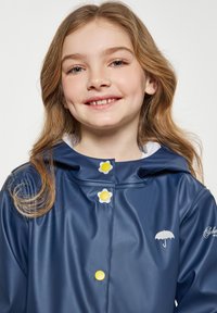 Mornarsko modra vodoodporna dežna bunda s kapuco, ki ima rumene cvetlične gumbe in majhen bel dežnik na prsih. Gladka tekstura.