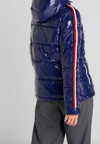 Giubbotto imbottito navy con finitura lucida, design trapuntato, cappuccio e strisce verticali rosse e bianche a contrasto lungo le maniche.