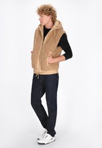 Gilet beige peloso con cappuccio, chiusura frontale con zip e due tasche; indossato sopra una maglietta nera a maniche lunghe e jeans scuri. Sneakers bianche.