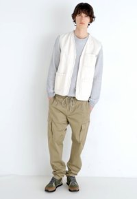Vesta albă fără mâneci peste o bluză cu mânecă lungă gri cu dungi, pantaloni cargo bej, pantofi verzi și maro. Design minimalist cu multiple buzunare.