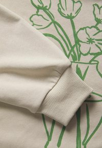 Beige sweatshirt sleeve with ribbed cuff resting on fabric printed with green floral line art of tulips and poppies.
