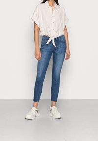 Camisa de botones de manga corta color beige claro anudada a la cintura, combinada con jeans ajustados azules y zapatillas blancas con detalles de estampado animal.