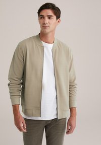 Chaqueta bomber beige clara con patrón texturizado, puños acanalados y cremallera frontal. Llevada sobre una camiseta blanca y combinada con pantalones verdes oliva.