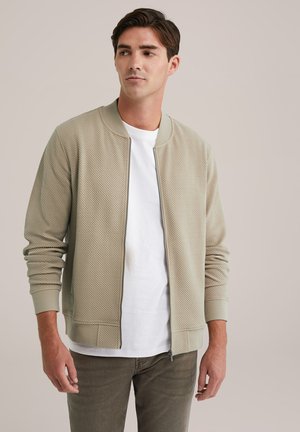 Light beige bomber jacket with textured pattern, ribbed cuffs, and a front zipper. Worn over a white T-shirt and paired with olive green pants.