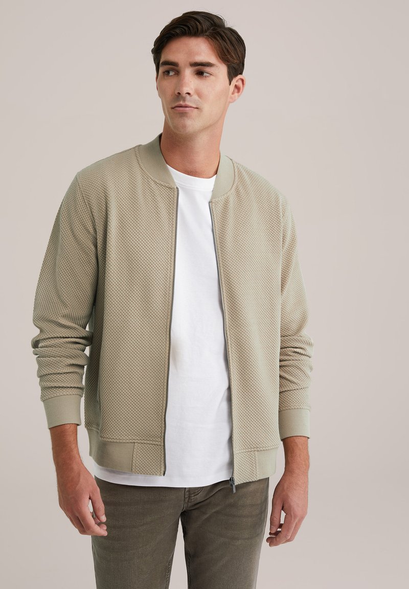 Chaqueta bomber beige clara con patrón texturizado, puños acanalados y cremallera frontal. Llevada sobre una camiseta blanca y combinada con pantalones verdes oliva.