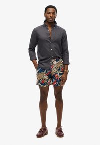Ciemnoszara koszula z guzikami, szorty w niebieskie i wielokolorowe wzory z tropikalnymi liśćmi, brązowe buty oraz zegarek na nadgarstku.
