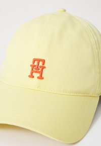 Baseball cap i lys gul stoff med en oransje brodert logo som har overlappende 'T' og 'H'. Glatt tekstur med sømdetaljer.