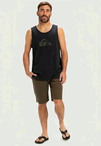 Camisola preta sem mangas com um logótipo verde, combinada com calções castanhos e chinelos pretos. Corte casual, tecido leve.