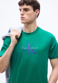 Νέος άνδρας που φοράει πράσινο t-shirt Calvin Klein, κρατώντας ένα ανοιχτό τζιν μπουφάν στον ώμο του, κοιτάζοντας προς τα αριστερά.