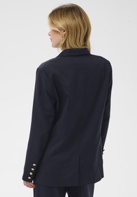 Blazer marine à coupe ajustée, avec un col à revers, des accents de boutons dorés sur les manches et un tissu lisse et texturé.