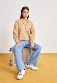 Camicia beige a maniche lunghe con tasca, jeans a gamba larga azzurri chiari e scarpe da ginnastica bianche. Modello indossa una collana a catena argentata, seduto su un baule metallico.