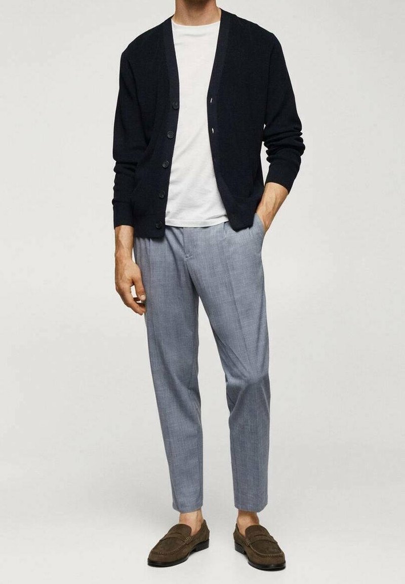 Homme portant un cardigan noir boutonné sur un t-shirt blanc, un pantalon gris clair et des mocassins marron, debout avec une main dans la poche.