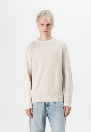 Mladý muž s krátkymi blond vlasmi nosí béžový pletený sveter a svetlomodré rifle, stojí pred jednoduchým bielym pozadím.