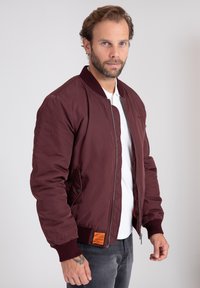 Giacca bomber bordeaux con polsini e colletto a costine, dotata di zip frontale e dettaglio etichetta arancione sul lato inferiore.