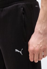 Κοντινό πλάνο ενός χεριού που ακουμπάει δίπλα σε μαύρο παντελόνι φόρμας Puma με πλάγιες πάνελ από δίχτυ και εμφανές μικρό τσεπάκι με φερμουάρ.