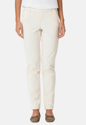 Femeie purtând pantaloni bej conici, bluză albă și balerini maro, stând într-un cadru simplu, cu fundal deschis la culoare.