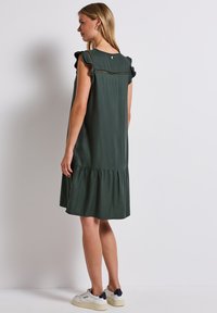 Zielona sukienka z krótkim rękawem, ozdobiona falbanami na ramionach oraz warstwowym dołem. Wykonana z miękkiego materiału o gładkiej fakturze, z dekoltem w kształcie łezki z tyłu.