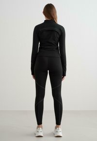 Černý sportovní top s dlouhými rukávy a vysokým límcem, v kombinaci s černými legínami, které těsně přiléhají a mají šedé švy.
