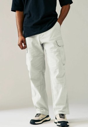 Homme portant un pantalon cargo gris clair avec des poches à rabat et des baskets noires et beiges, debout devant un fond neutre.