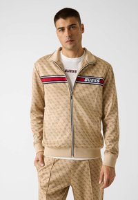 Giacca da pista beige con motivo logo, zip frontale e strisce contrastanti rosse e nere. Abbinate a pantaloni coordinati. Tessuto strutturato.