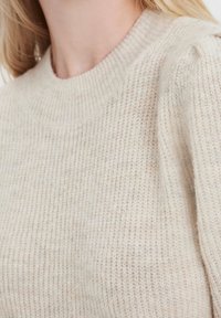 Ljust beige stickad tröja med rund halsringning, texturerad yta och subtilt mönster, som har lätt puffiga axeldetaljer.