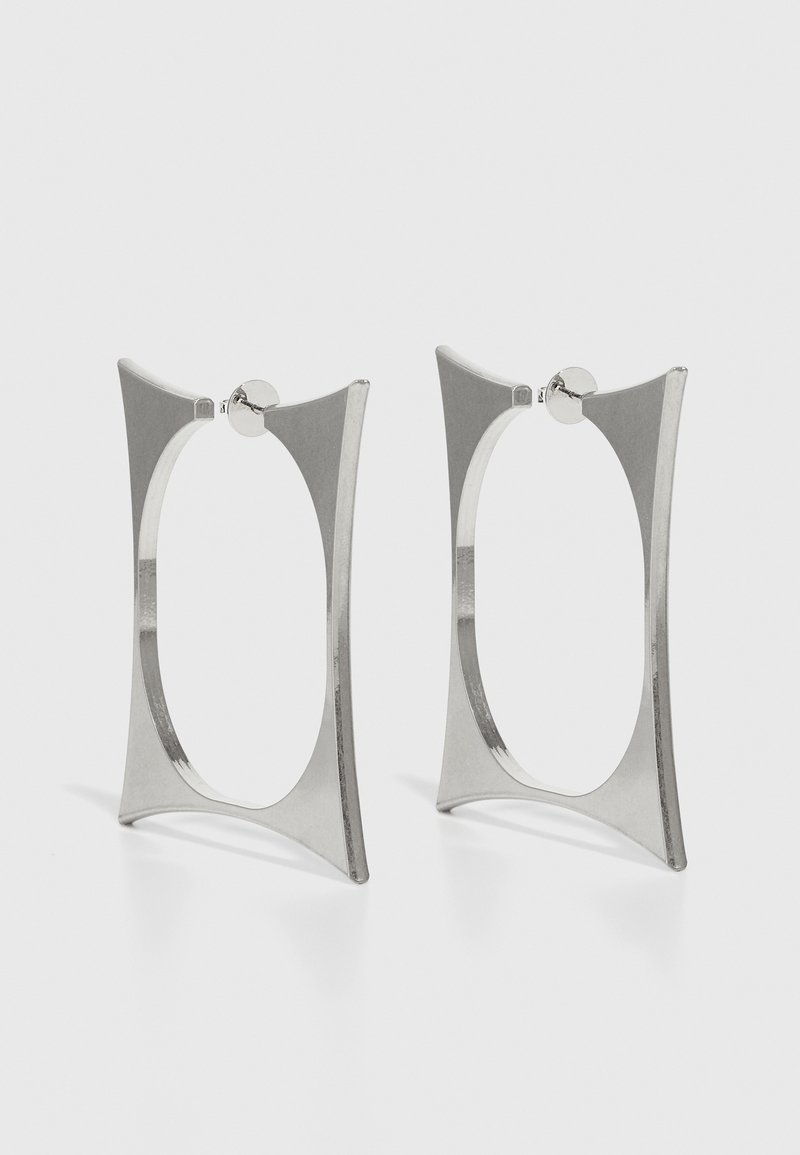 Srebrne obruče za uši s geometrijskim dizajnom, koje sadrže izdužene, kutne oblike i polirani metalni završni sloj. Zaključavanje na vrhu.