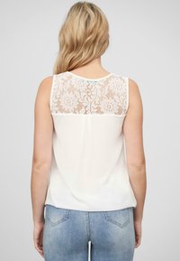 Haut sans manches blanc avec un dos en dentelle présentant des motifs floraux. L'avant est uni et le tissu est lisse et léger.