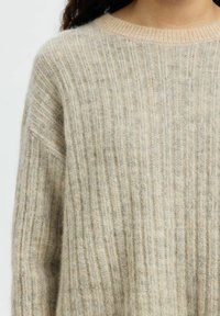 Gros plan sur une personne portant un pull en tricot côtelé beige avec une texture douce et pelucheuse et un col rond.