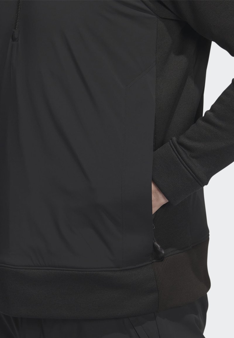 Person in schwarzer Jacke mit Hand in der seitlichen Reißverschlusstasche, die Texturen des Stoffes sowie Nahtdetails an Ärmel und Saum zeigt.