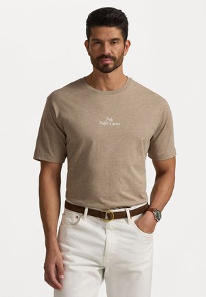 T-shirt à manches courtes beige avec "Polo Ralph Lauren" brodé en blanc. Porté avec une ceinture marron et un jean blanc ; texture lisse, coupe décontractée.