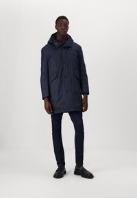 Navy blue nylon parka with a hood, featuring front buttons, two side pockets, and a slightly elongated shape. Styled over dark jeans and shoes.