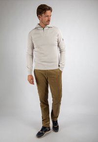 Man in beige geribbelde trui met ritshals, bruine corduroy broek en zwarte sneakers, staand tegen een effen achtergrond.