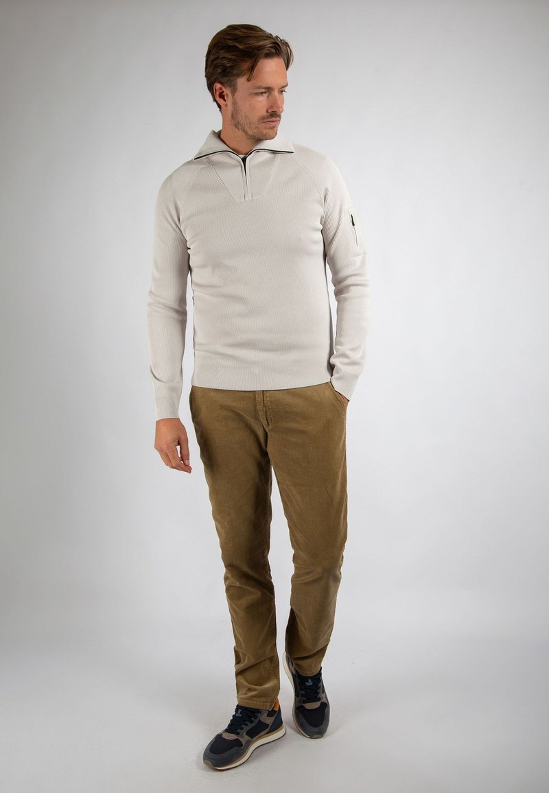Man in beige geribbelde trui met ritshals, bruine corduroy broek en zwarte sneakers, staand tegen een effen achtergrond.