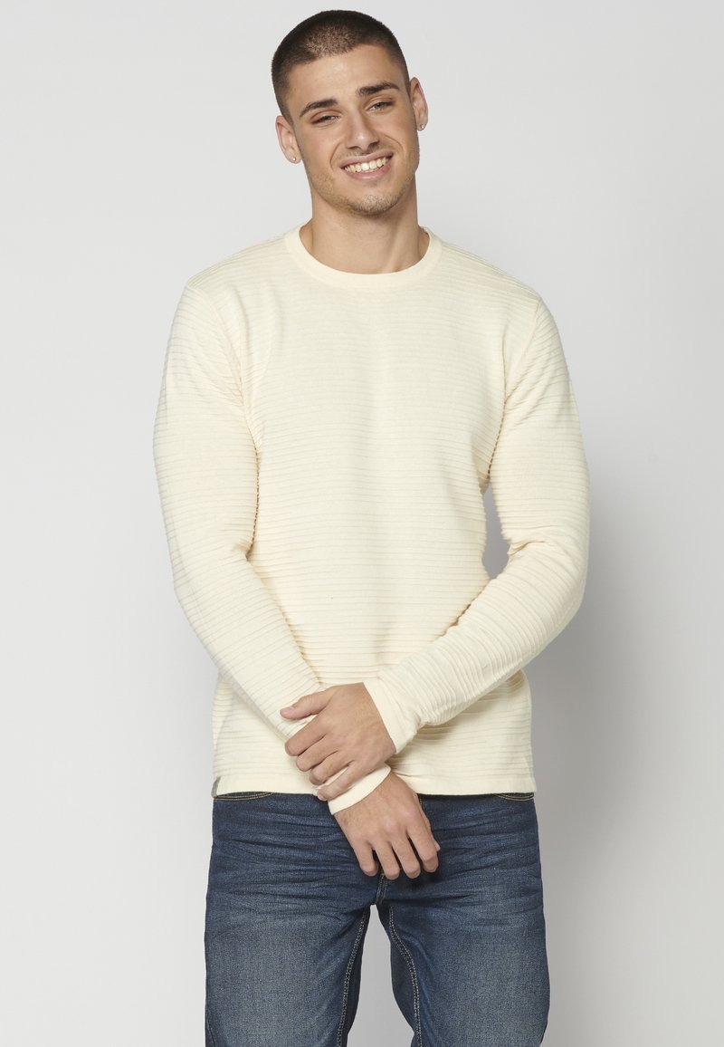 Hellbeige, langärmliger Pullover mit strukturiertem Rippdesign. Kombiniert mit dunkelblauen Jeans, die Hände vor dem Körper verschränkt.