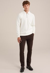 Hanorac alb cu fermoar și guler, purtat împreună cu pantaloni maro închis și pantofi negri. Textură netedă, design mulat. Aspect simplu, modern.