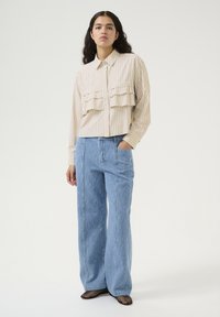Blusa beige a righe con due tasche frontali arricciate, abbinata a pantaloni in denim leggeri e larghi di colore blu chiaro con righe verticali e tasche frontali.