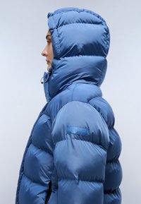 Giacca piumino blu con colletto alto e cappuccio. Presenta una texture lucida e liscia, un motivo a righe orizzontali e una toppa con logo sulla manica.