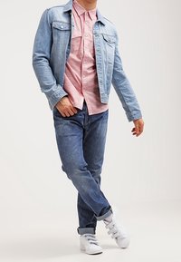 Giacca in denim azzurro chiaro sopra una camicia rosa con colletto, abbinata a jeans blu e sneakers bianche, che mettono in mostra un look rilassato e uno stile informale.