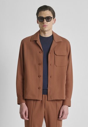 Man met een bruine jasje met knopen en klepzak op de borst, bijpassende broek, donkerblauw overhemd en zwarte zonnebril tegen een effen achtergrond.