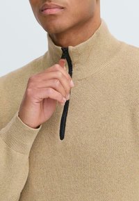 Beige gebreide trui met een hoge kraag en zwart ritsdetail, voorzien van een subtiele textuur en geribbelde manchetten. Hand past de rits aan.