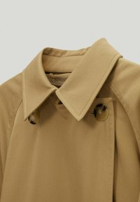 Trench-coat kaki avec un col classique, équipé de deux grandes fermetures à boutons et d'une texture lisse et tissée. Aucune motif ou accent visible.