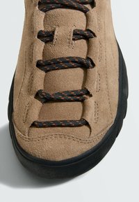 Gros plan sur le bout d'une basket en daim beige avec des lacets ronds noirs et orange et une épaisse semelle en caoutchouc noir sur un fond clair.
