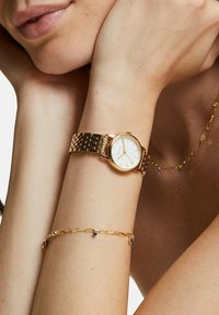 Gul armbandsur med en rund vit urtavla och en länkad kedja, parat med ett delikat gult armband med små blå detaljer.