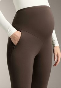 Hnědé těhotenské legíny s vysokým pasem, hladkým materiálem a bočními kapsami. Materiál vypadá pružně a dobře obepíná postavu.