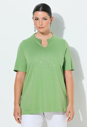 Kvinde med mørkt hår iført en grøn kortærmet top med rhinsten lodrette striber, iført store hængende øreringe, foran en helt hvid baggrund.