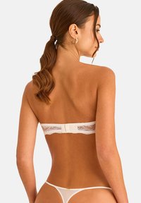 Femme aux cheveux bruns attachés en queue de cheval, portant un soutien-gorge en dentelle sans bretelles et un string en résille, vue de dos sur un fond uni.